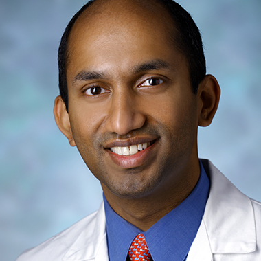 Chetan Bettegowda, in a formal portrait, wearing a white lab coat, blue shirt and red tie.